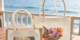 Deux sacs en jute personnalisés, grand et petit modèle, posés sur une table en bois avec vue sur la mer.